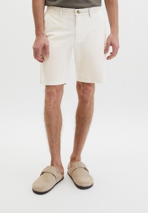 Man die beige korte broek tot aan de knieën en beige instapschoenen draagt, staand tegen een effen achtergrond.