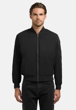 Mężczyzna ubrany w czarną kurtkę bomber z zamkiem błyskawicznym, z prążkowanymi mankietami i kołnierzem, w ciemnych dżinsach, stojący na jasnym, jednolitym tle.