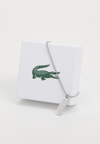 Collier en chaîne argenté avec une étiquette rectangulaire gravée "LACOSTE," présenté avec une boîte texturée blanche arborant un logo de crocodile vert.