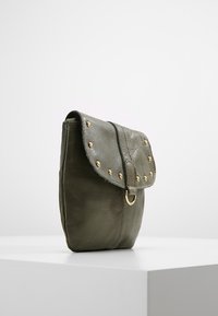 Borsa a mano in pelle verde oliva con chiusura a patta, borchie dorate lungo il bordo e un accento metallico circolare. Superficie strutturata, forma compatta.
