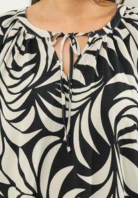 Bluse i sort og creme med et blomsterprint, blødt stof, rund halsudskæring med bindebånd og elastiske ærmekanter.