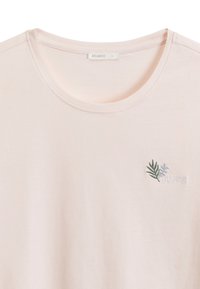 Jasnoróżowa koszulka krótki rękaw z okrągłym dekoltem i subtelnym zielonym haftem w kształcie liścia po lewej stronie klatki piersiowej.