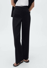 Pantaloni neri a vita alta con un design a gamba dritta, caratterizzati da una texture liscia e linee pulite. Abbinati a una blusa bianca e sandali.