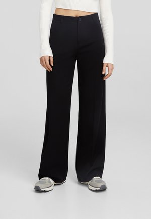Femme portant un pantalon large noir et des baskets grises, assortis à un pull blanc court à manches longues, debout sur un fond uni.