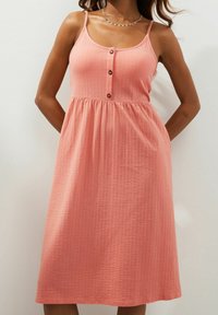 Vestido sin mangas de color rosa coral con tela texturizada, detalles de botones y falda acampanada, con corsé ajustado y bolsillos laterales.