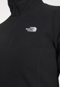 Detailní záběr na černou fleecovou bundu s polovičním zipem a vyšitým bílým logem "The North Face" na prsou.