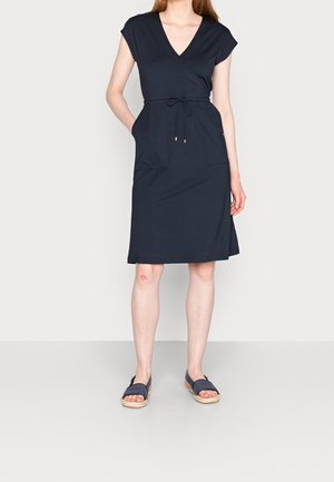 Kobieta w granatowej sukience kopertowej do kolan z krótkimi rękawami, przednimi kieszeniami i wiązaniem w talii, zestawiona z granatowymi sandałami z odkrytymi palcami.