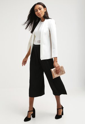 White tailored blazer, black culottes, and a fitted white top with cutouts. Holding a metallic gold clutch. Black high-heeled shoes.