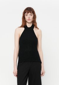 Kvinne med rødt hår som har på seg en svart ermeløs topper med teksturerte mønstre og svarte bukser mot en enkel hvit bakgrunn.