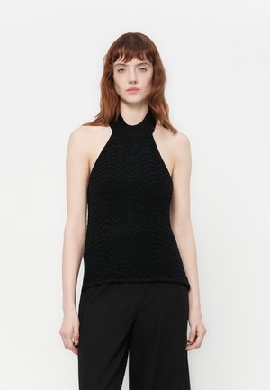Femme avec des cheveux roux portant un top halter sans manches noir avec des motifs texturés et un pantalon noir, sur un fond blanc uni.
