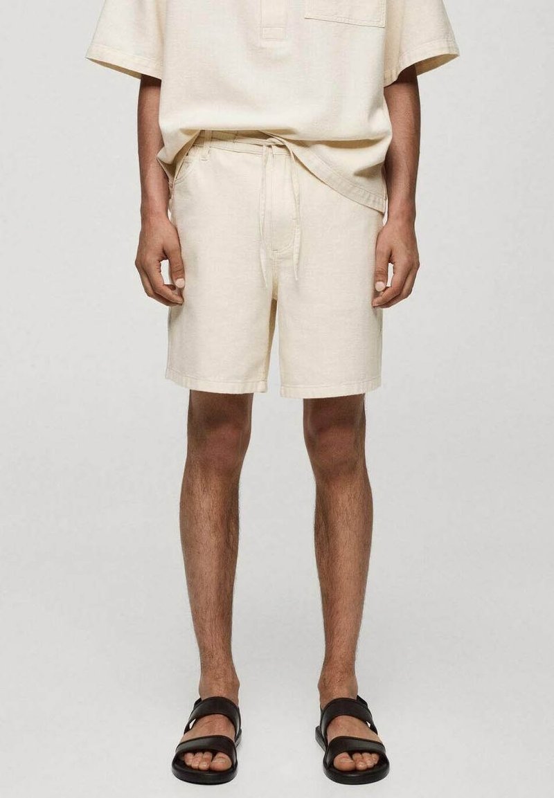 Homme portant un short beige à cordon de serrage, une chemise beige assortie et des sandales noires à double bride, debout devant un fond uni.