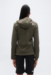 Λαδί πράσινο μπουφάν με κουκούλα, εφαρμοστό σχέδιο, κατασκευασμένο από λείο ύφασμα, με λογότυπο στο πάνω μέρος της πλάτης, σε συνδυασμό με μαύρο σορτς.