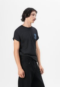 Black cotton t-shirt with rolled sleeves and blue text that reads “RIDE WITH RA” on the left chest, paired with black denim pants.