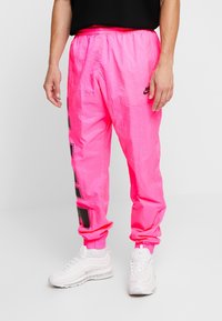 Man wearing bright pink Nike track pants with black logo and white Nike sneakers, standing on white floor against plain background.