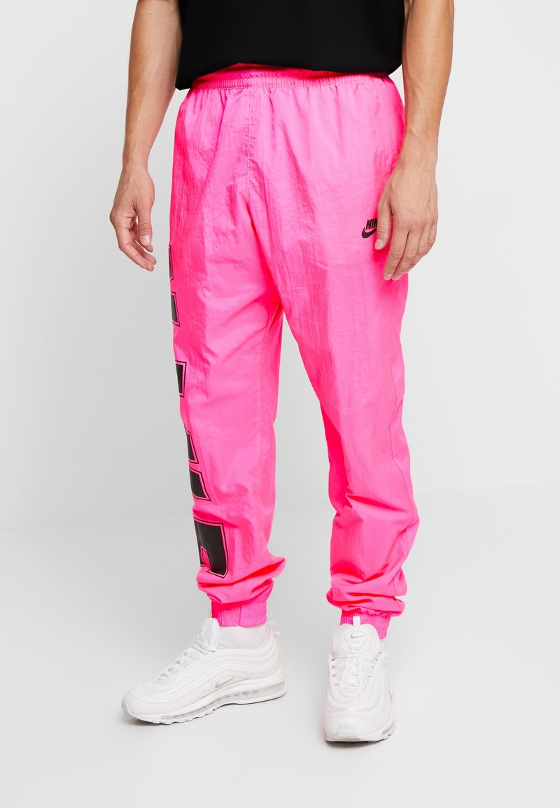 Man wearing bright pink Nike track pants with black logo and white Nike sneakers, standing on white floor against plain background.