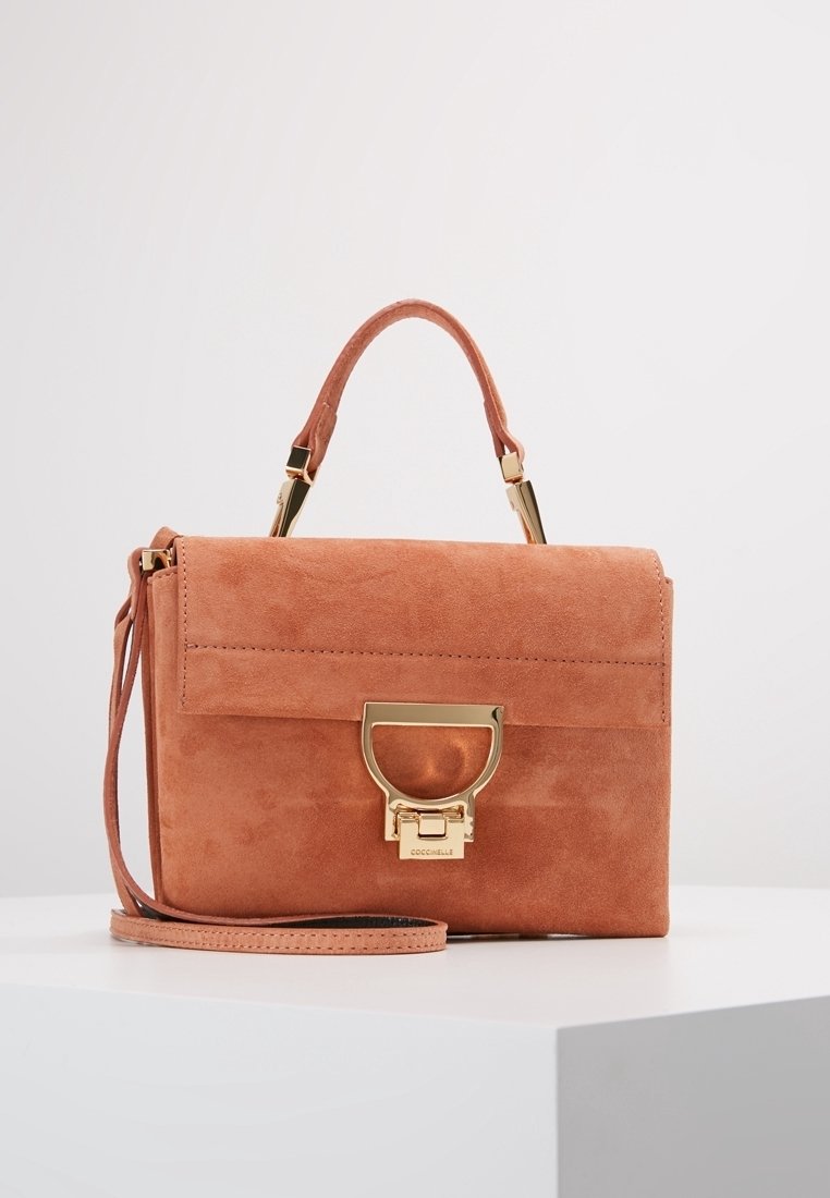 Prostokątna torebka z zamszu w kolorze przybrudzonego różu, z zapięciem w złotym odcieniu, górnym uchwytem i odczepianym paskiem na ramię, umieszczona na białej powierzchni.