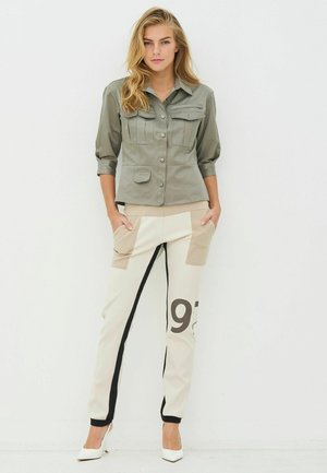 Olivengrønn jakke med knappelukking og to brystlommer, kombinert med beige og kremfargede joggebukser med grafiske tall og sidestreker.