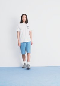 Hvit t-skjorte med grafikk på brystet, blå denimshorts og grå joggesko. Står på lyseblått gulv mot hvit vegg. Langt hår.