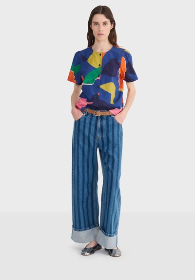 Frau trägt eine bunte Bluse mit abstraktem Muster und Knöpfen, weit geschnittene Jeans mit gestreiften Umschlägen und braunem Gürtel sowie schwarze Ballettschuhe, stehend.