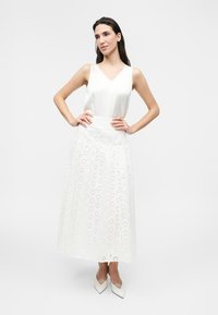 Γυναίκα που στέκεται με τα χέρια στη μέση, φοράει αμάνικη λευκή μπλούζα, μακριά λευκή φούστα με κομμένο λουλουδάτο σχέδιο και λευκά μυτερά παπούτσια.