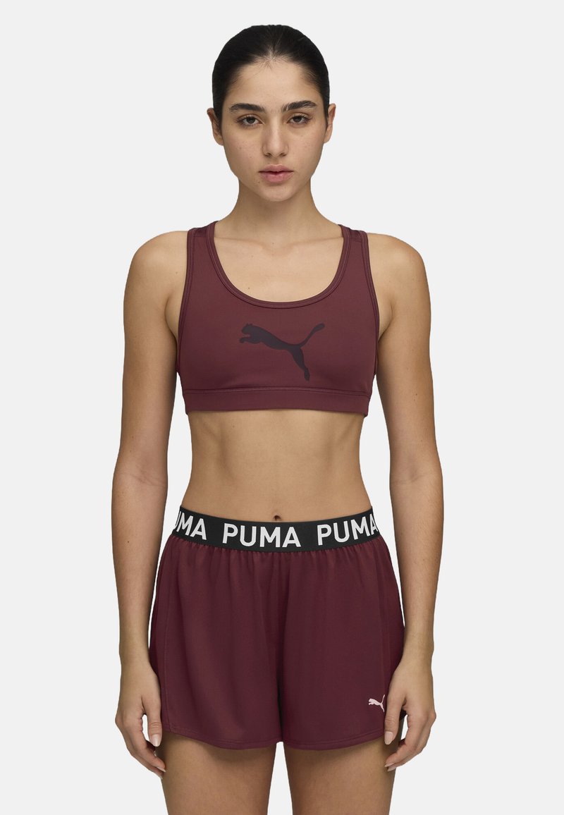 Bordeaux sportski grudnjak s crnim logotipom, uparen s odgovarajućim bordeaux kratkim hlačama koje imaju crni pojas s bijelim "PUMA" tekstom. Glatka tekstura.