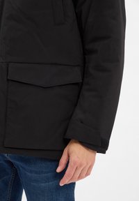 Zwarte geïsoleerde jas met een ritssluiting, een klepzak en gestructureerde manchetten. Beschikt over een stijlvolle, gladde buitenkant en een getailleerde pasvorm.