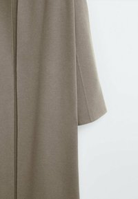 Manteau long en laine couleur taupe à manches longues suspendu sur un fond blanc uni, montrant la couture latérale et le détail de la manche.