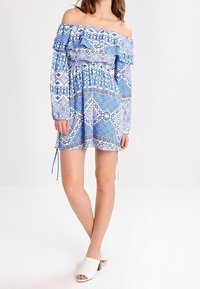 Abito blu a maniche lunghe, scollato sulle spalle, con motivi a fantasia e rouches, vita elastica e lacci decorativi. Abbinato a mule bianche con punta aperta.