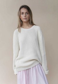 Flauschiger, creamfarbener Strickpullover mit weitem Ausschnitt und geripptem Muster, kombiniert mit einem pastellrosa Rock, der einen geschichteten Look kreiert.