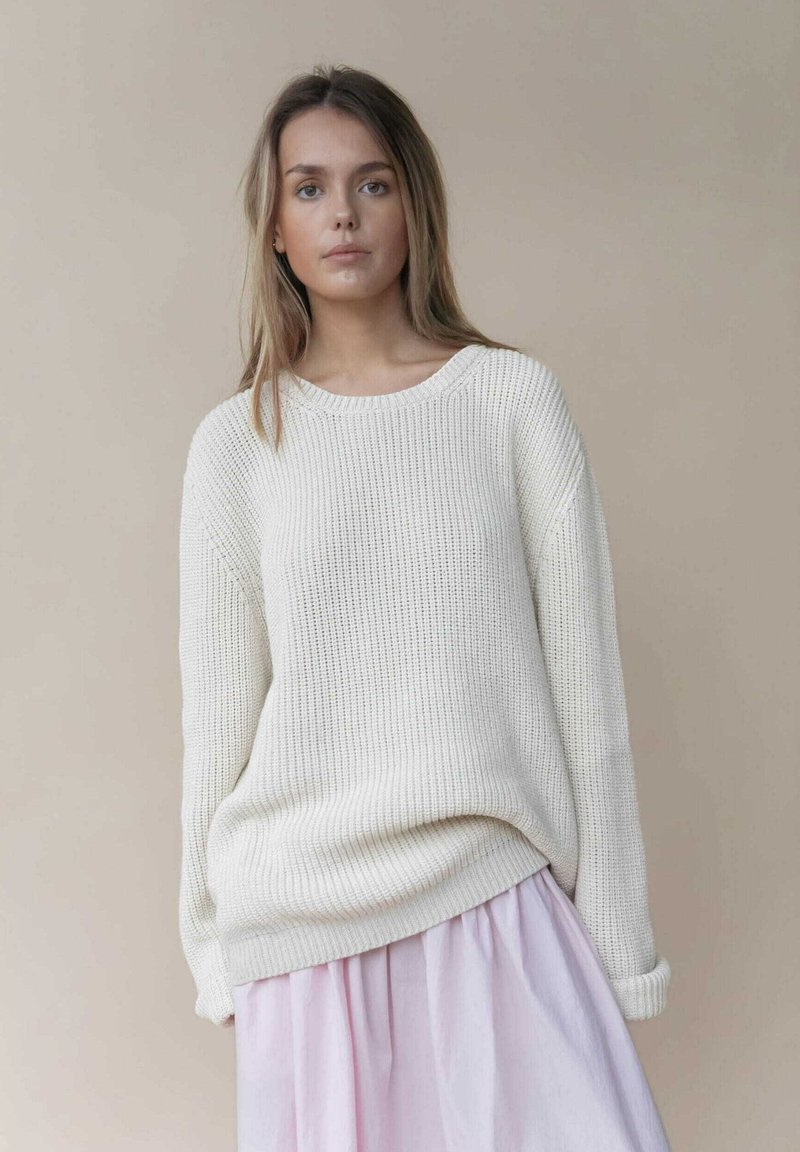 Flauschiger, creamfarbener Strickpullover mit weitem Ausschnitt und geripptem Muster, kombiniert mit einem pastellrosa Rock, der einen geschichteten Look kreiert.