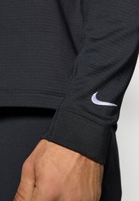 Μαύρη αθλητική μπλούζα με μακριά μανίκια και λευκό λογότυπο Nike στον καρπό, φοριέται από άτομο με ορατό χέρι και καρπό.