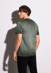 Zelené tričko s krátkým rukávem, které má texturovaný gradientní vzor a kulatý výstřih, v kombinaci s černými kalhotami. Obsahuje prošívané okraje.