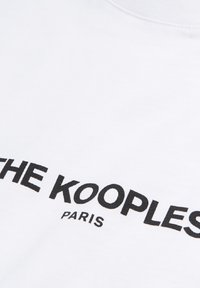 Camiseta blanca de algodón con texto impreso en negro "THE KOOPLES PARIS." Textura suave, diseño de cuello redondo y sin patrones adicionales.
