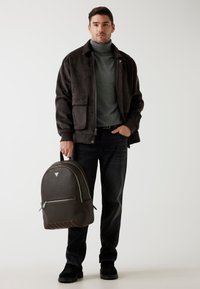 Mørkebrun rygsæk med struktureret materiale, sølvhardware og frontlomme. Modellen har en matchende turtleneck og jakke på, samt sorte jeans.