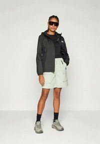 Kvinde iført sort vindjakke, sort t-shirt, lyse cargo-shorts, vandresko, sorte sokker og store sorte solbriller, som står foran en hvid baggrund.