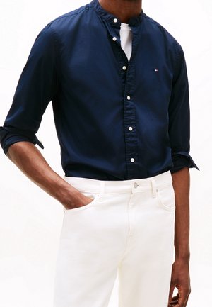 Homme portant une chemise bleu marine boutonnée avec les manches retroussées et un pantalon blanc, main dans la poche devant un fond uni.