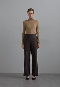 Beige eng anliegendes Langarmoberteil mit hohem Kragen, kombiniert mit hoch sitzenden, dunkelbraunen, weit geschnittenen Hosen. Schwarze spitz zulaufende Ballerinas runden das Outfit ab.