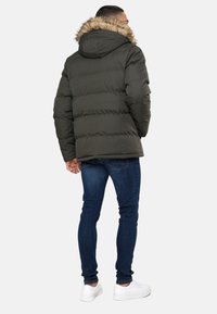 Grüne Pufferjacke mit einem mit Fell gefütterten Kapuze, ausgestattet mit einem gesteppten Design, kombiniert mit dunkelblauen Jeans und weißen Sneakers.