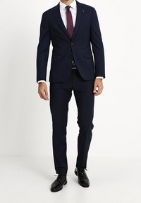 Uomo che indossa un completo scuro in tessuto aderente, camicia bianca, cravatta bordeaux a fantasia, cintura nera e scarpe eleganti nere lucide.