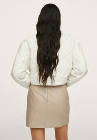 Giacca imbottita bianca e trapuntata con maniche larghe, indossata sopra una mini gonna in pelle beige, vista da dietro con lunghi capelli scuri visibili.