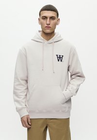Light pink hoodie with a front pocket, drawstring hood, and a dark blue logo on the left chest, paired with tan pants.
