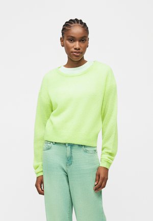 Γυναίκα με πλεξούδες, που φοράει ένα πουλοβεράκι σε λαϊμ πράσινο χρώμα και ανοιχτοπράσινο τζιν ψηλόμεσο, στέκεται μπροστά σε απλό λευκό φόντο.