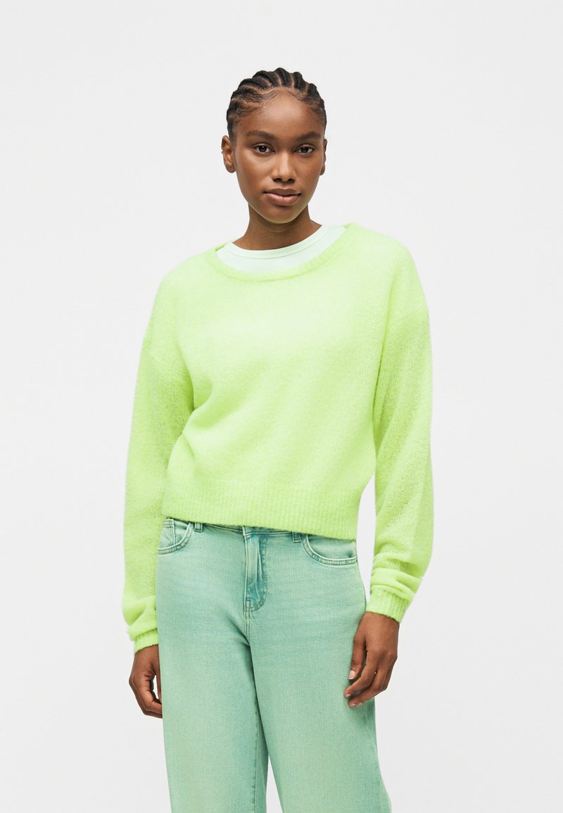 Γυναίκα με πλεξούδες, που φοράει ένα πουλοβεράκι σε λαϊμ πράσινο χρώμα και ανοιχτοπράσινο τζιν ψηλόμεσο, στέκεται μπροστά σε απλό λευκό φόντο.