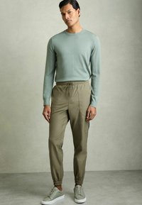 Pullover verde deschis cu mâneci lungi, asociat cu pantaloni jogger bej cu talie elastică și buzunare laterale, completat cu adidași gri deschis.