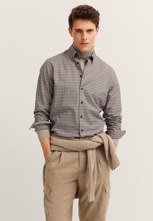 Mann in einem braun karierten Hemd, das über einem beigen Rollkragenpullover getragen wird, mit beigen Hosen und einem beigen Pullover, der um die Taille gebunden ist.