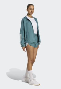 Teal athletic jacket with hood, white inner shirt, matching teal shorts, white socks, and light sneakers with textured accents. Three stripes on sleeves.