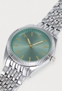 Montre ronde avec un cadran vert menthe, des index horaires dorés et un bracelet en maille en acier inoxydable argenté. Comprend une lunette texturée et une couronne.
