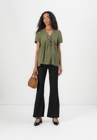 Haut enveloppant à manches courtes vert olive avec une taille froncée, associé à un pantalon évasé noir présentant un motif texturé subtil. Sac à main marron et escarpins noirs.