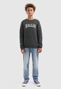 Grijze gebreide trui met "BALLIN" in witte letters, gecombineerd met lichtblauwe jeans en blauwe sneakers. Natuurlijke vezelstructuur zichtbaar.
