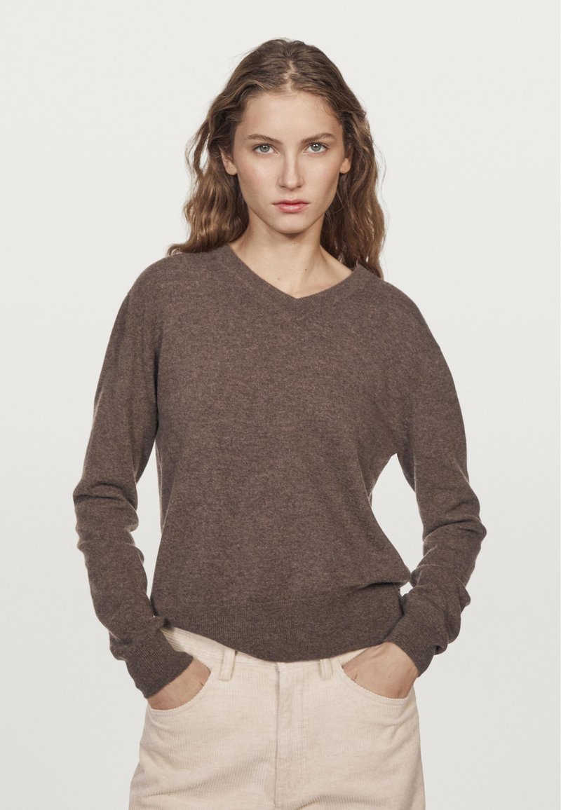 Brauner V-Ausschnitt-Pullover aus weichem Strickstoff. Lange Ärmel mit Rippbündchen. Kombiniert mit hellen Cordhosen. Einfaches Design, minimalistischer Stil.